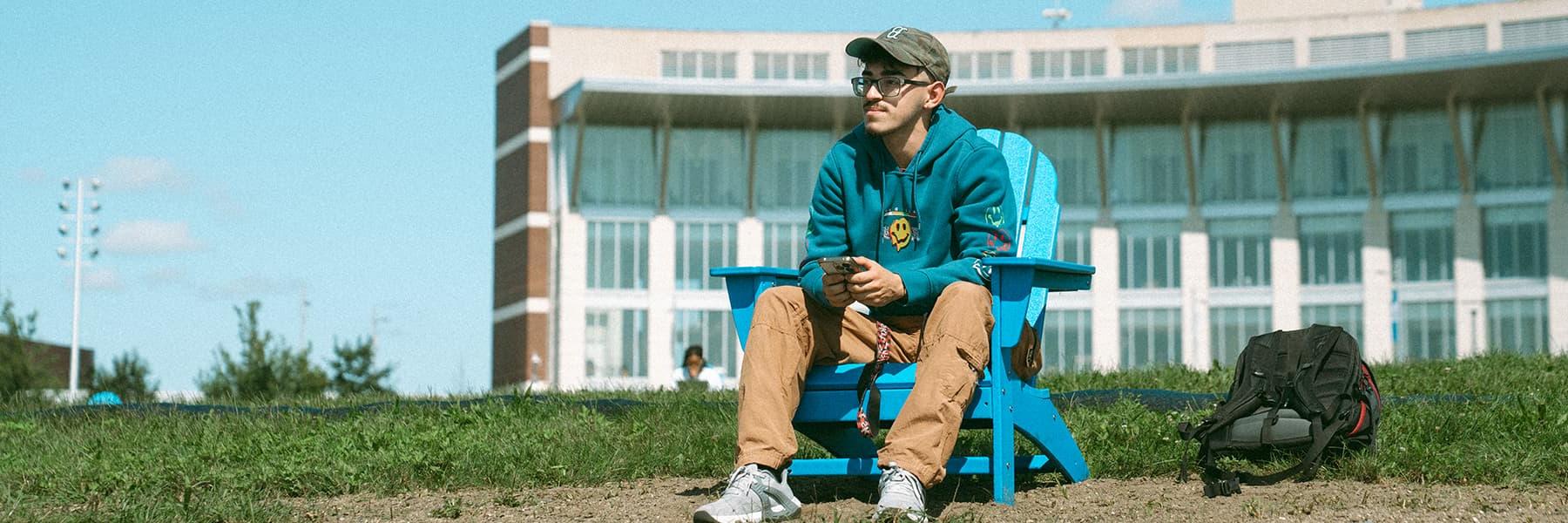 Student sits in blue chair on campus center lawn with cellphone in hand.